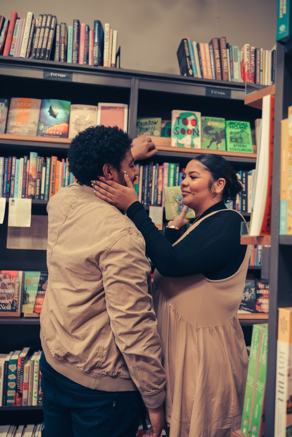 A couple embraces lovingly between bookshelves in a cozy bookstore setting.