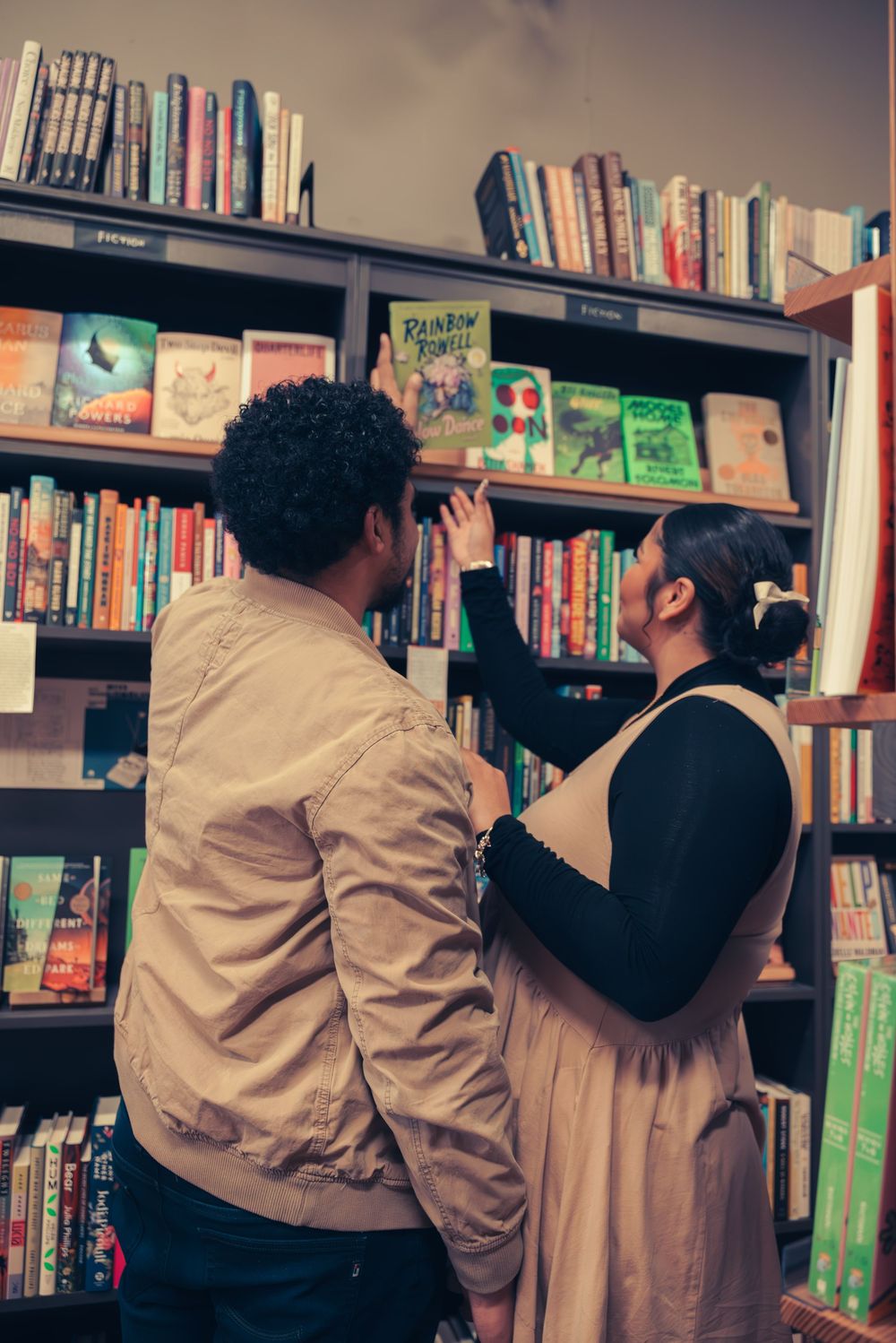 Two people browse books together on tall wooden bookshelves in a bookstore or library.