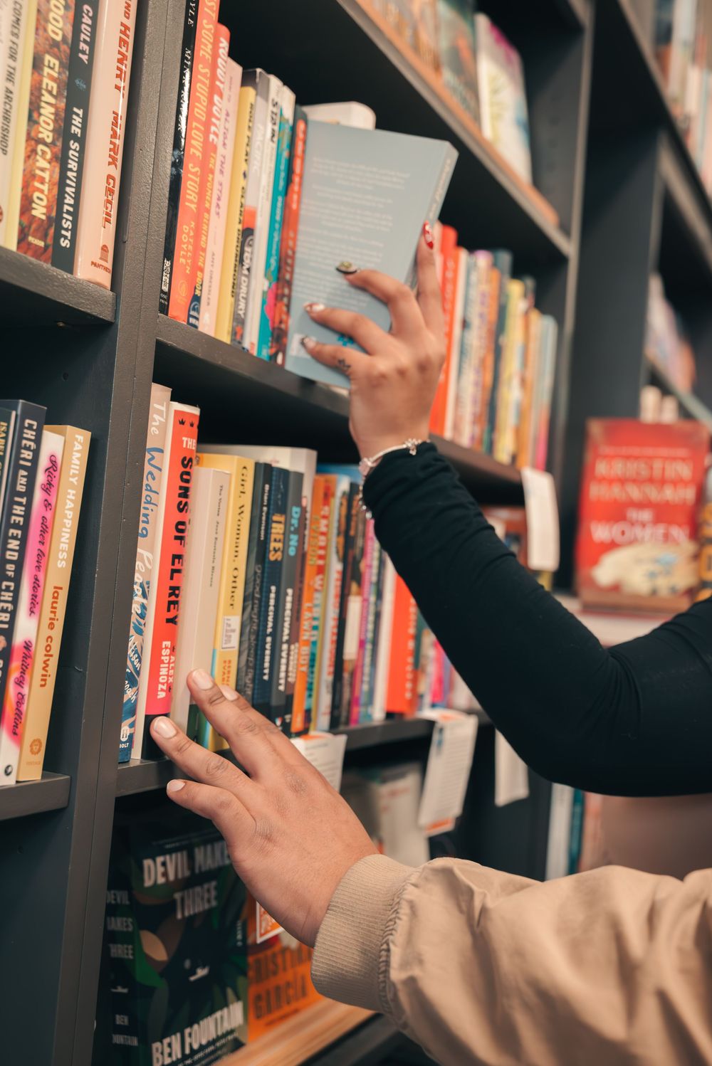 Hands reach for books on wooden library shelves filled with various colored book spines.