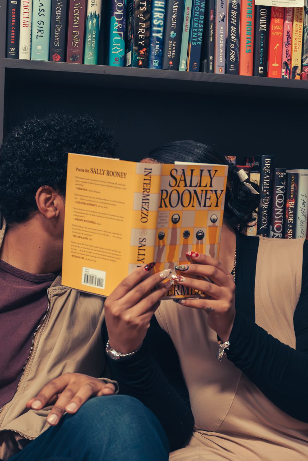 Two people hold books while sitting close together against a backdrop of library shelves.