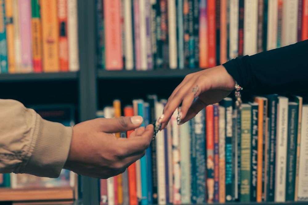 Two people exchanging books while standing in a library aisle with packed bookshelves.