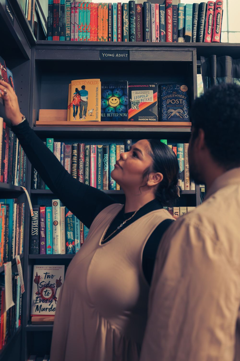 A person reaches up to select a book from a high shelf while another watches in a bookstore.