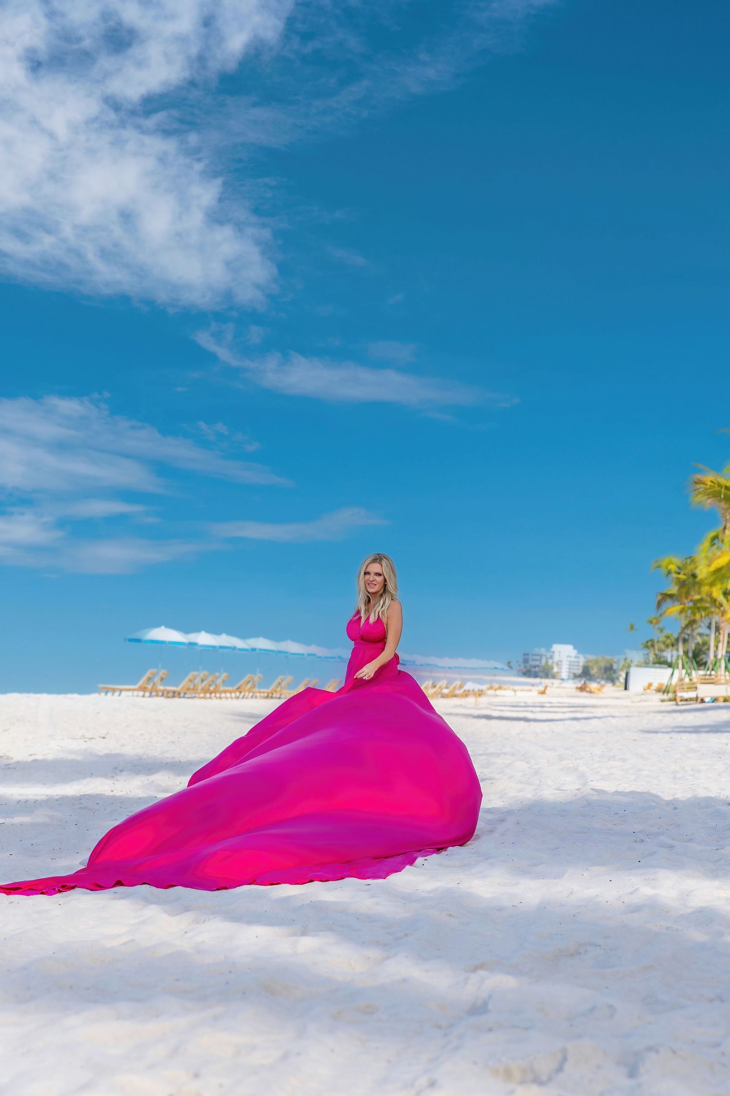 A vibrant pink fabric billows gracefully across pristine white sand beach against a bright blue tropical sky.