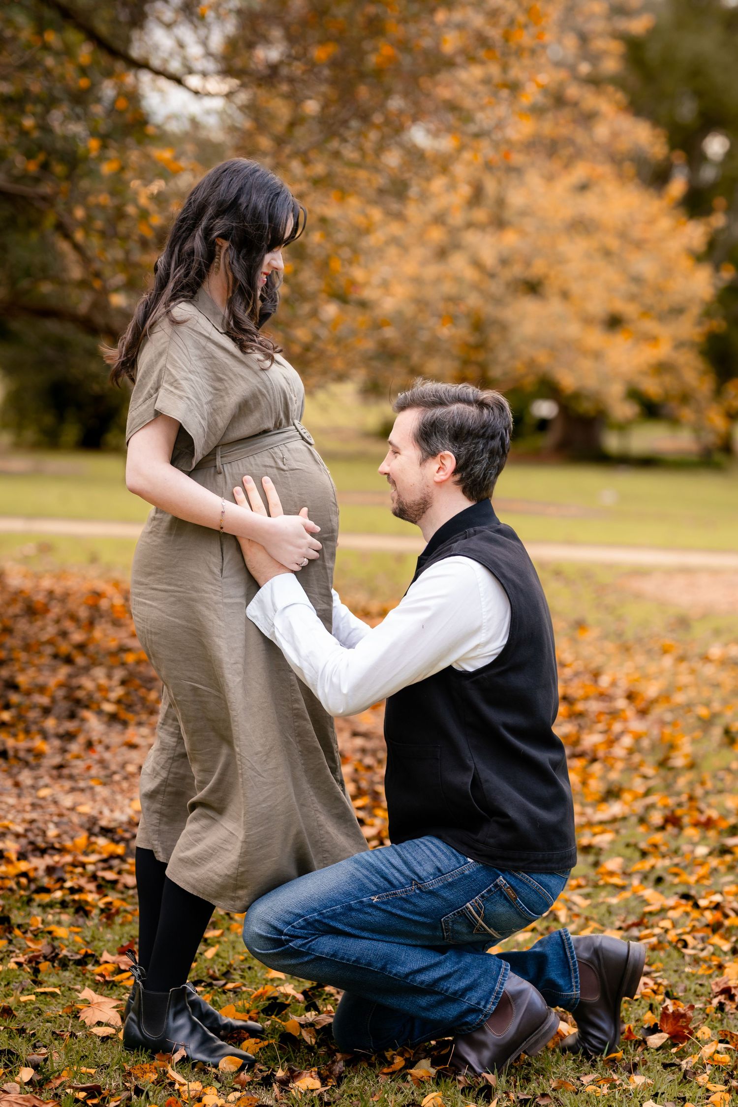 A dad-to-be kneeling with his hands on his partner's baby bump in an autumn park