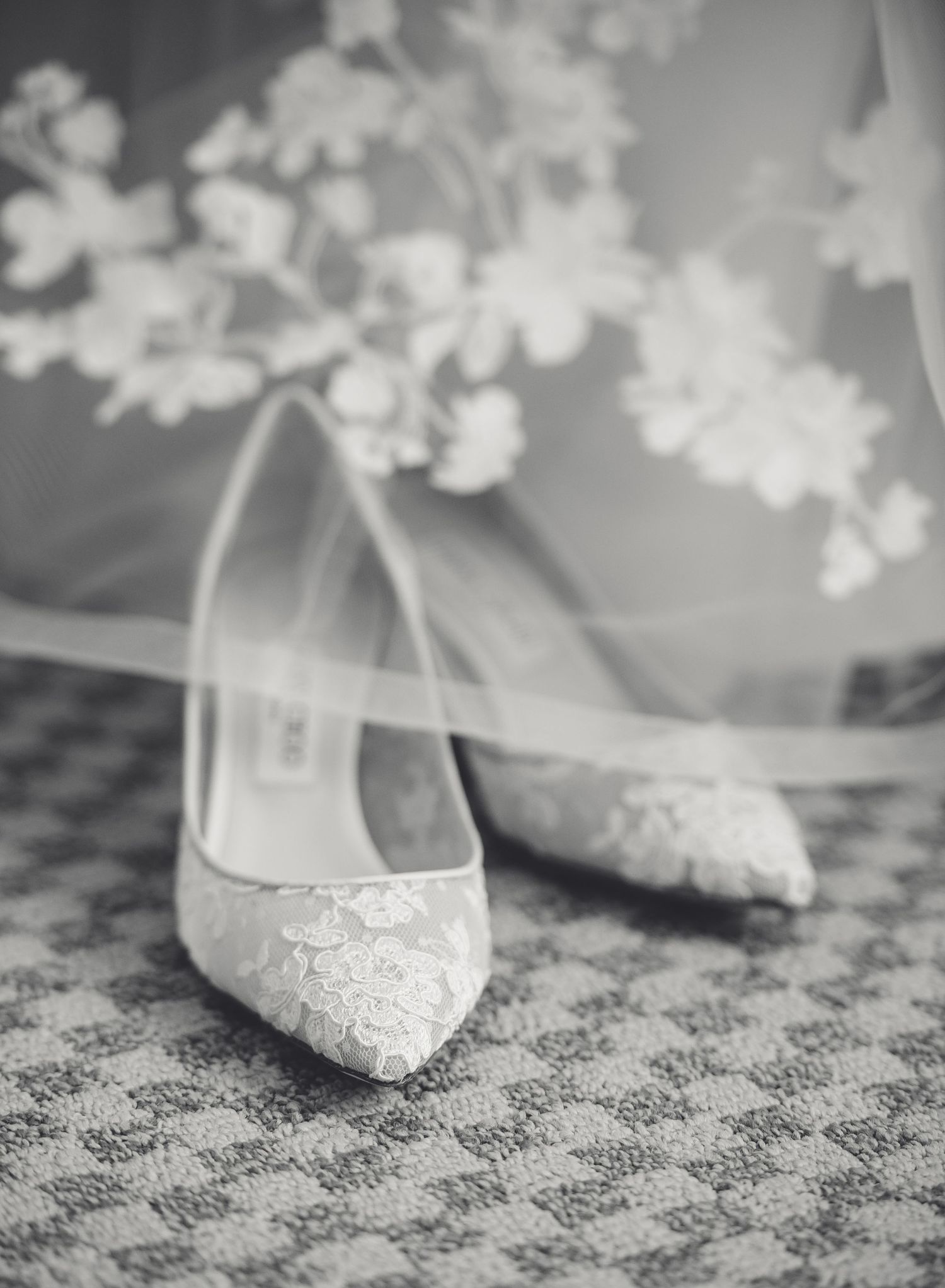 Black and white close-up photo of elegant lace bridal shoes on textured fabric with delicate floral details.
