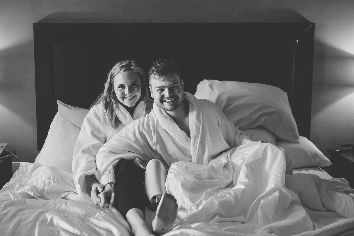 A black and white photograph of two people in white robes relaxing together on a bed with white linens.