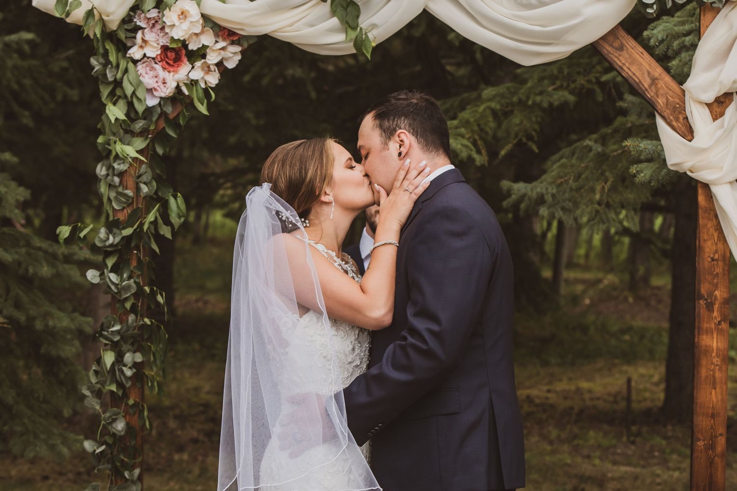 A romantic wedding couple shares an intimate moment under a wooden arch adorned with white and pink floral decorations.