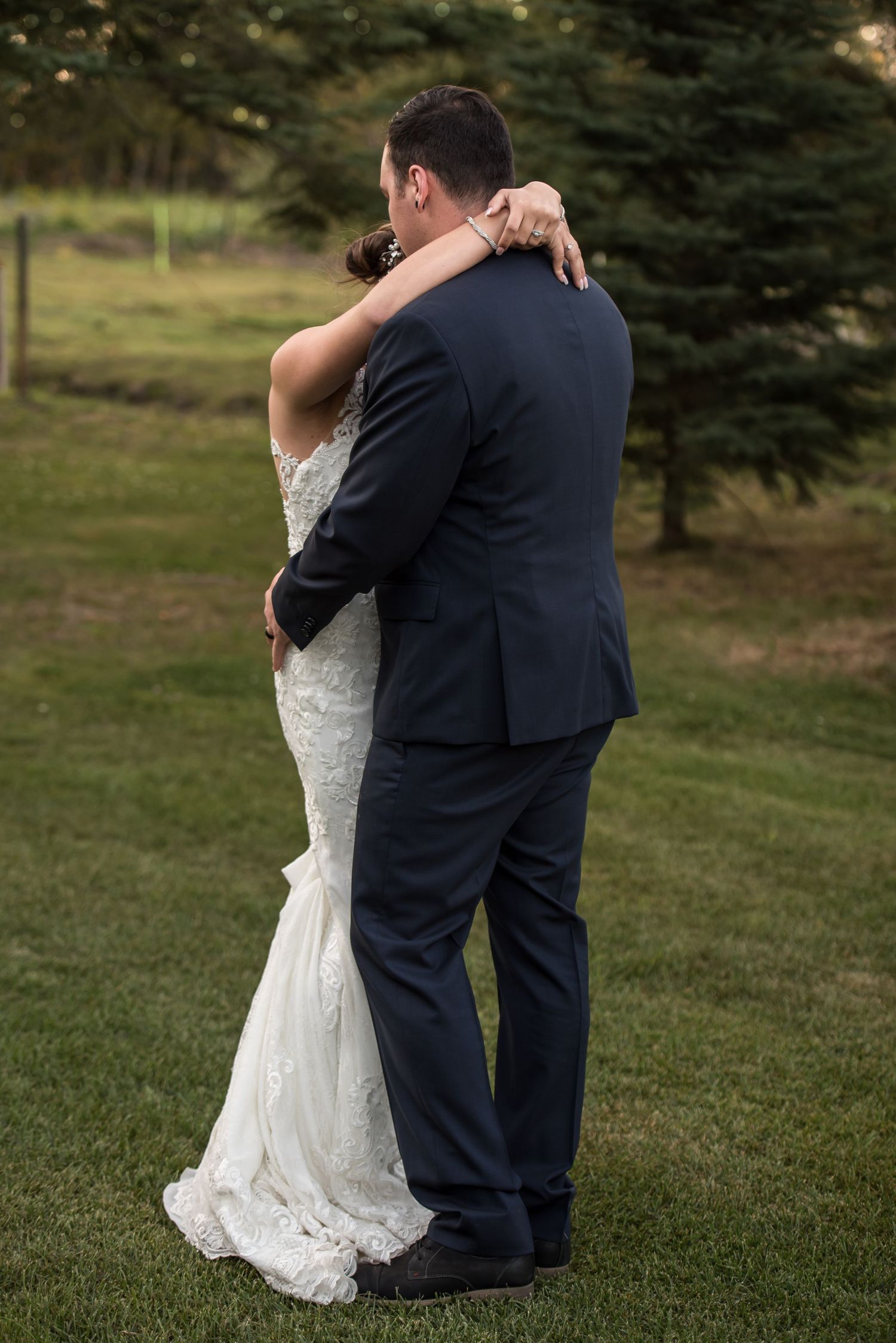 Couple in wedding attire embrace intimately outdoors with evergreen trees in the background.