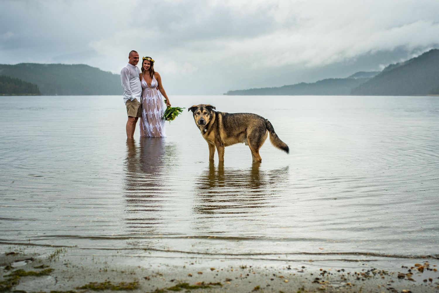 A couple and their German Shepherd dog stand in shallow water at a scenic mountain lake on a cloudy day.