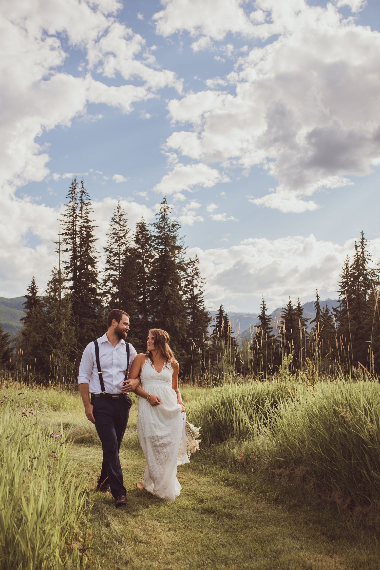 Couple in wedding attire walking through tall grass with evergreen trees and cloudy sky in mountain landscape.
