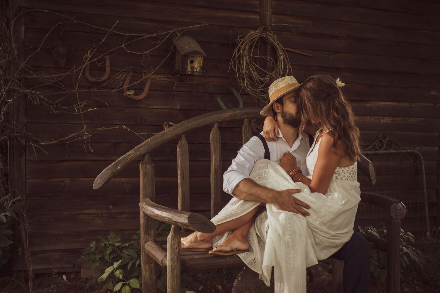 Romantic couple in white clothing embraces on rustic wooden porch with vintage decor and warm lighting.