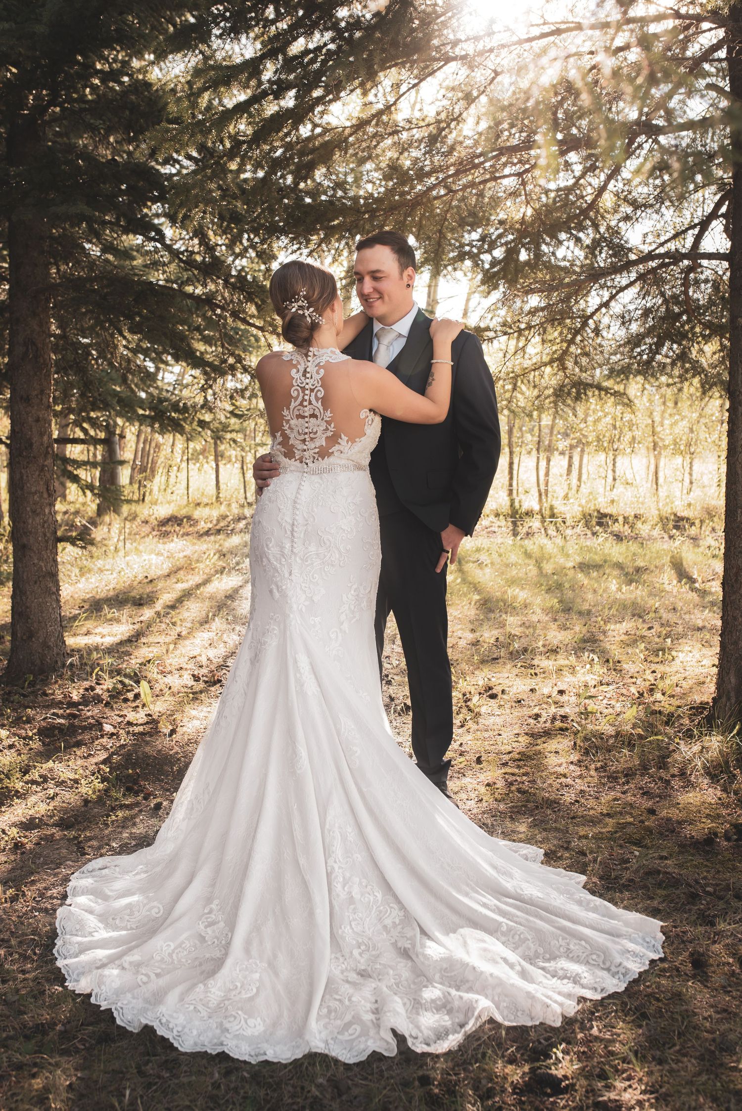 A romantic wedding couple dances in a sunlit forest with the bride's lace dress flowing elegantly around them.