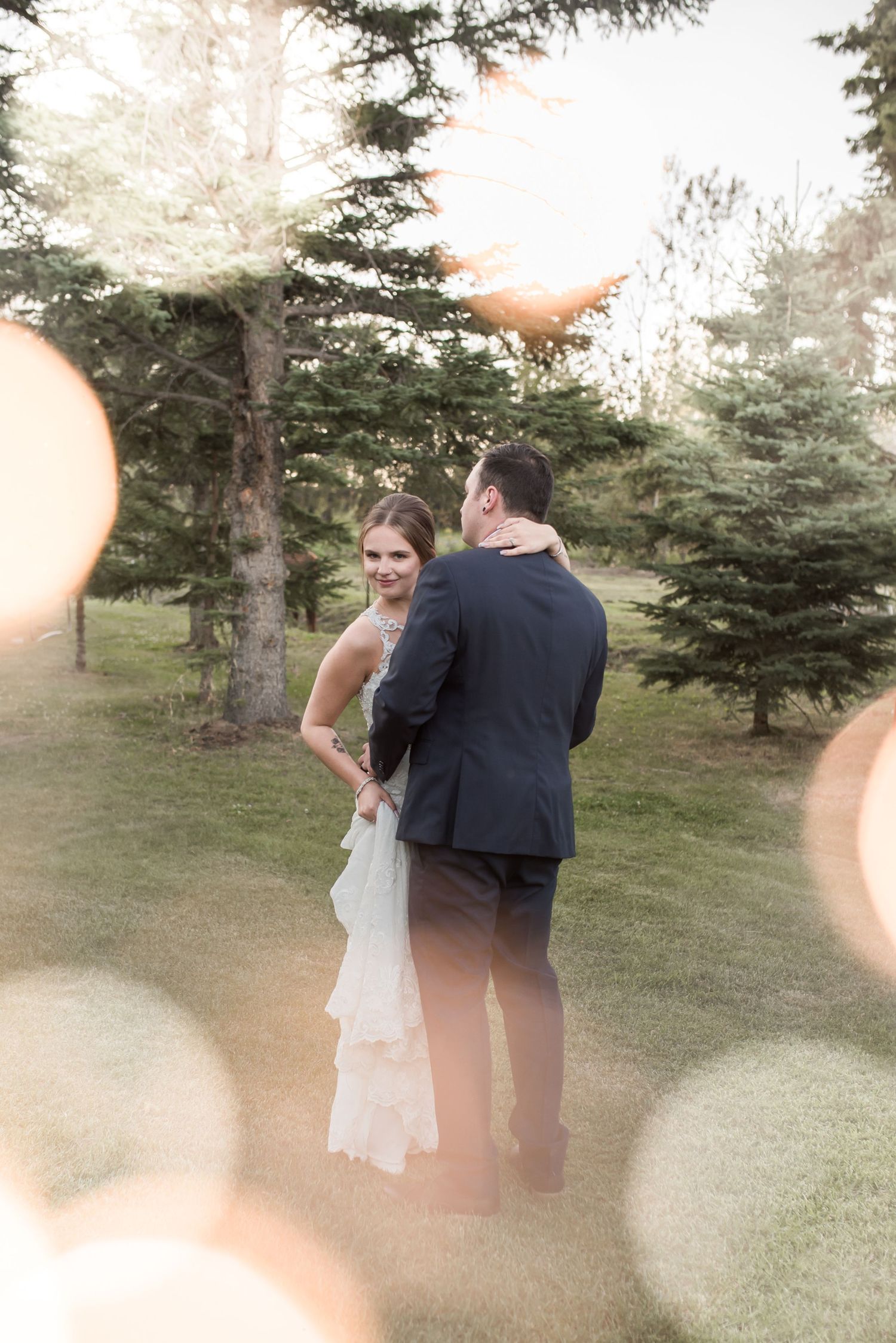 A romantic wedding couple shares an intimate moment outdoors among evergreen trees with dreamy bokeh lighting effects.