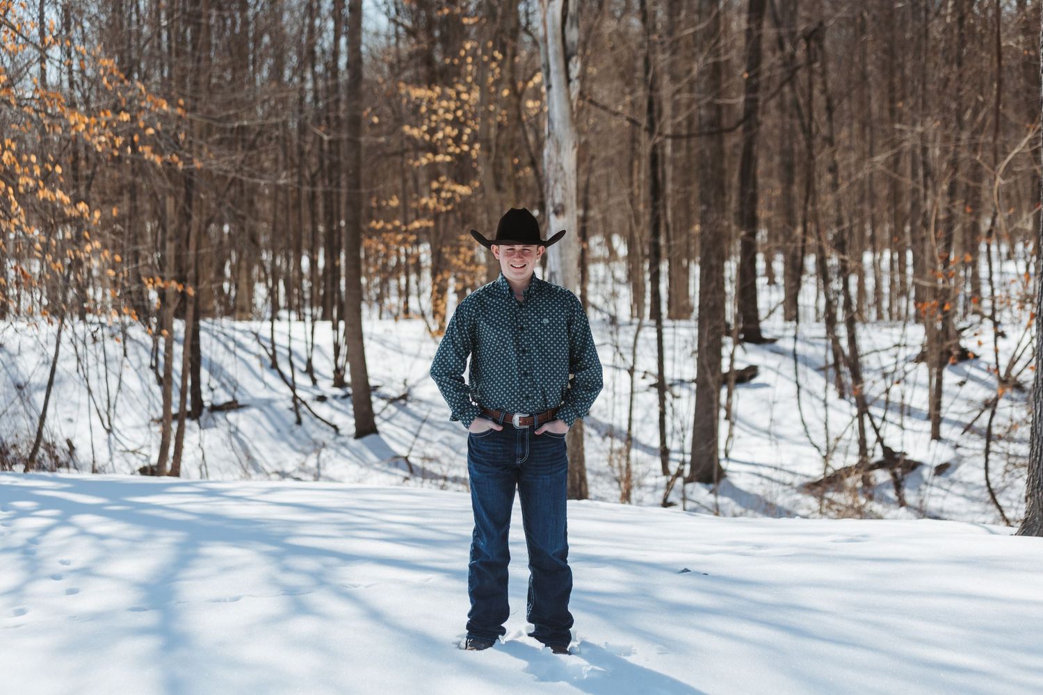 A person walks through a snowy winter forest landscape with bare trees and deep snow covering the ground.