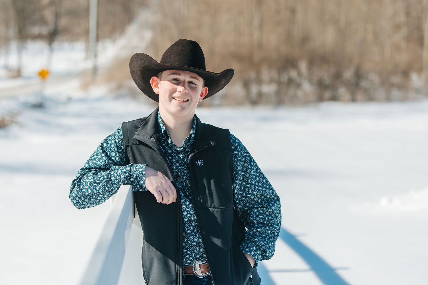A cowboy in western attire poses together in a snowy winter setting with mountains in the background.