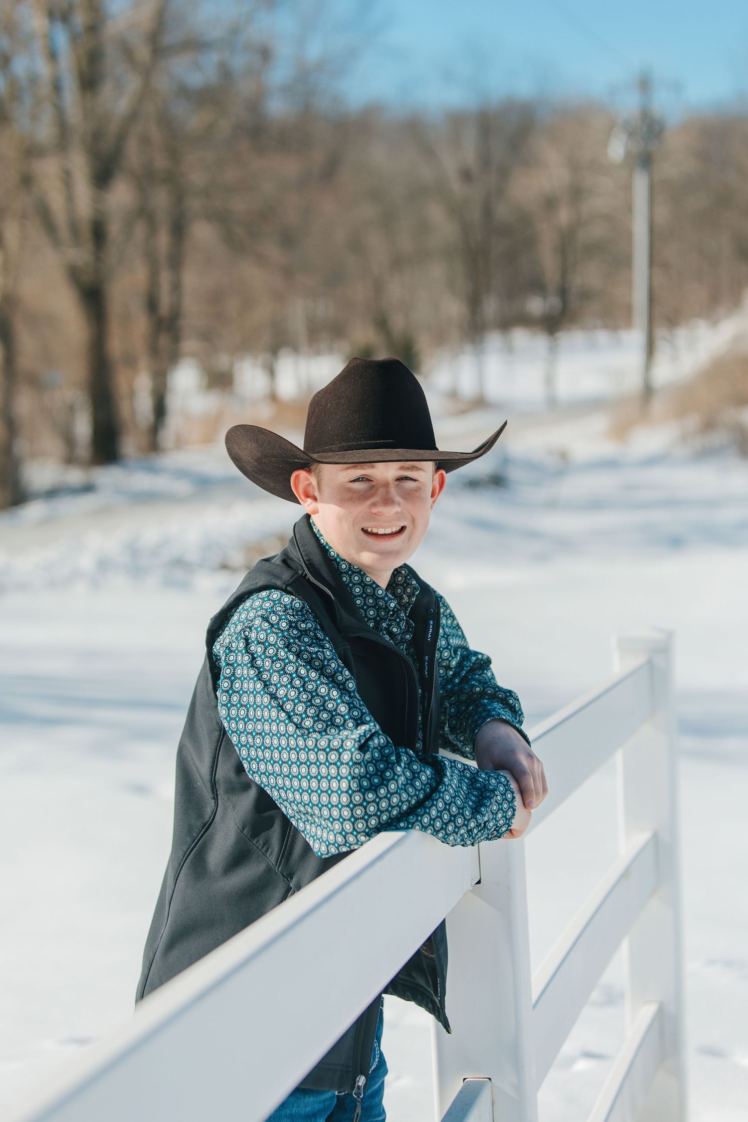 A person in a cowboy hat and scarf leans against a white fence in a snowy winter landscape.