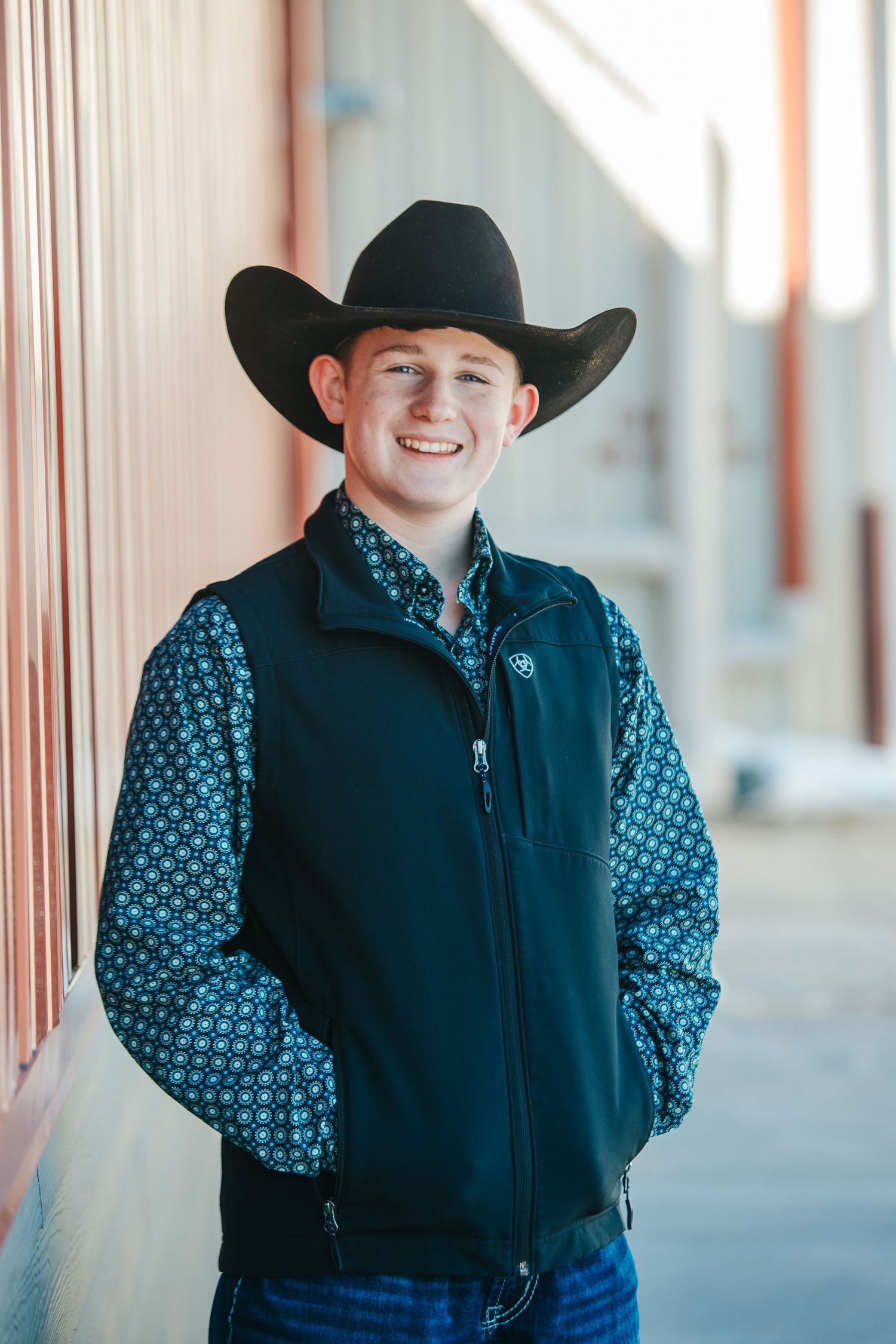 Young person in western attire with cowboy hat and patterned shirt poses against rustic wall.