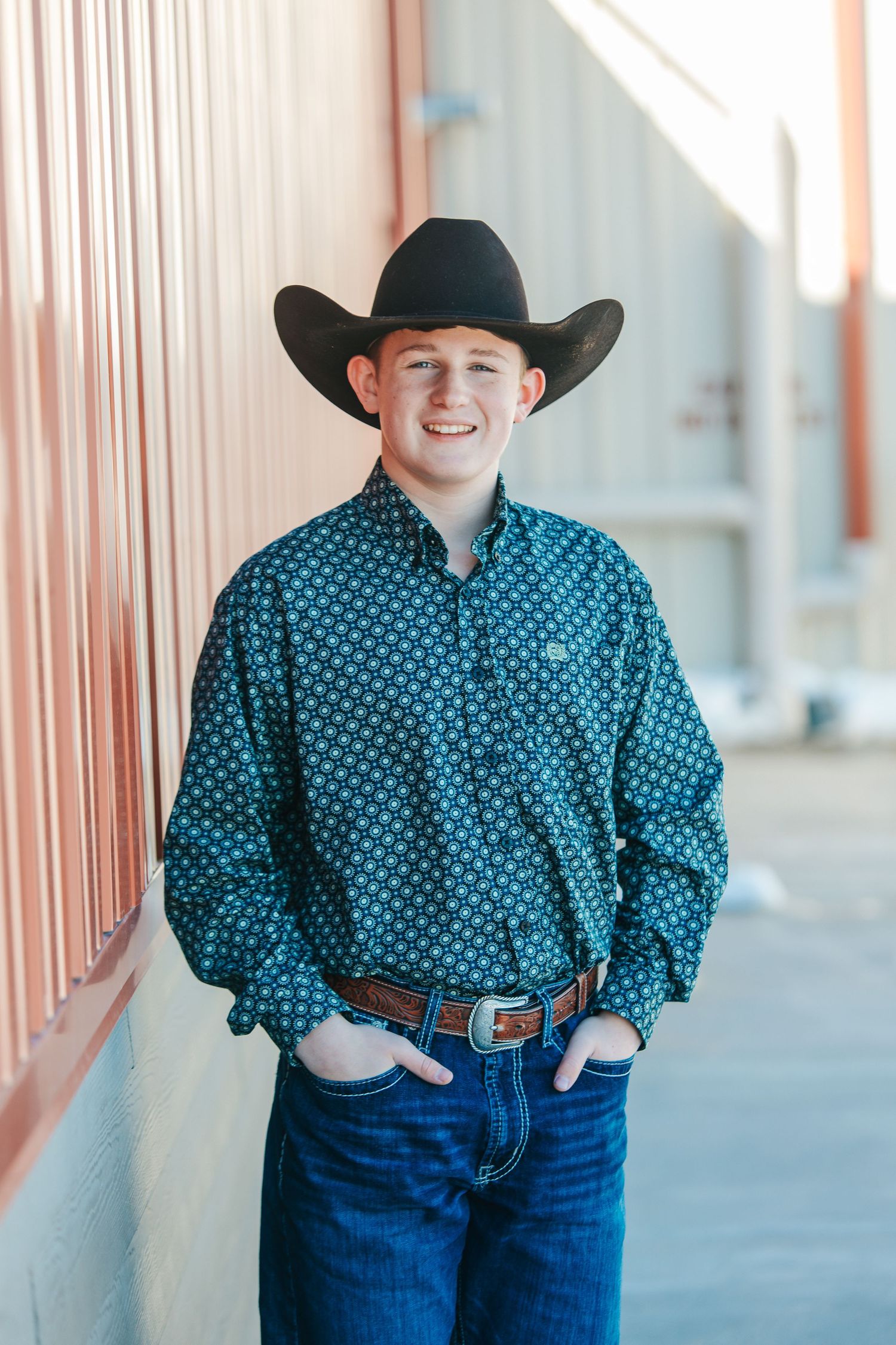 A person in a black cowboy hat and blue patterned button-down shirt poses against a wall.