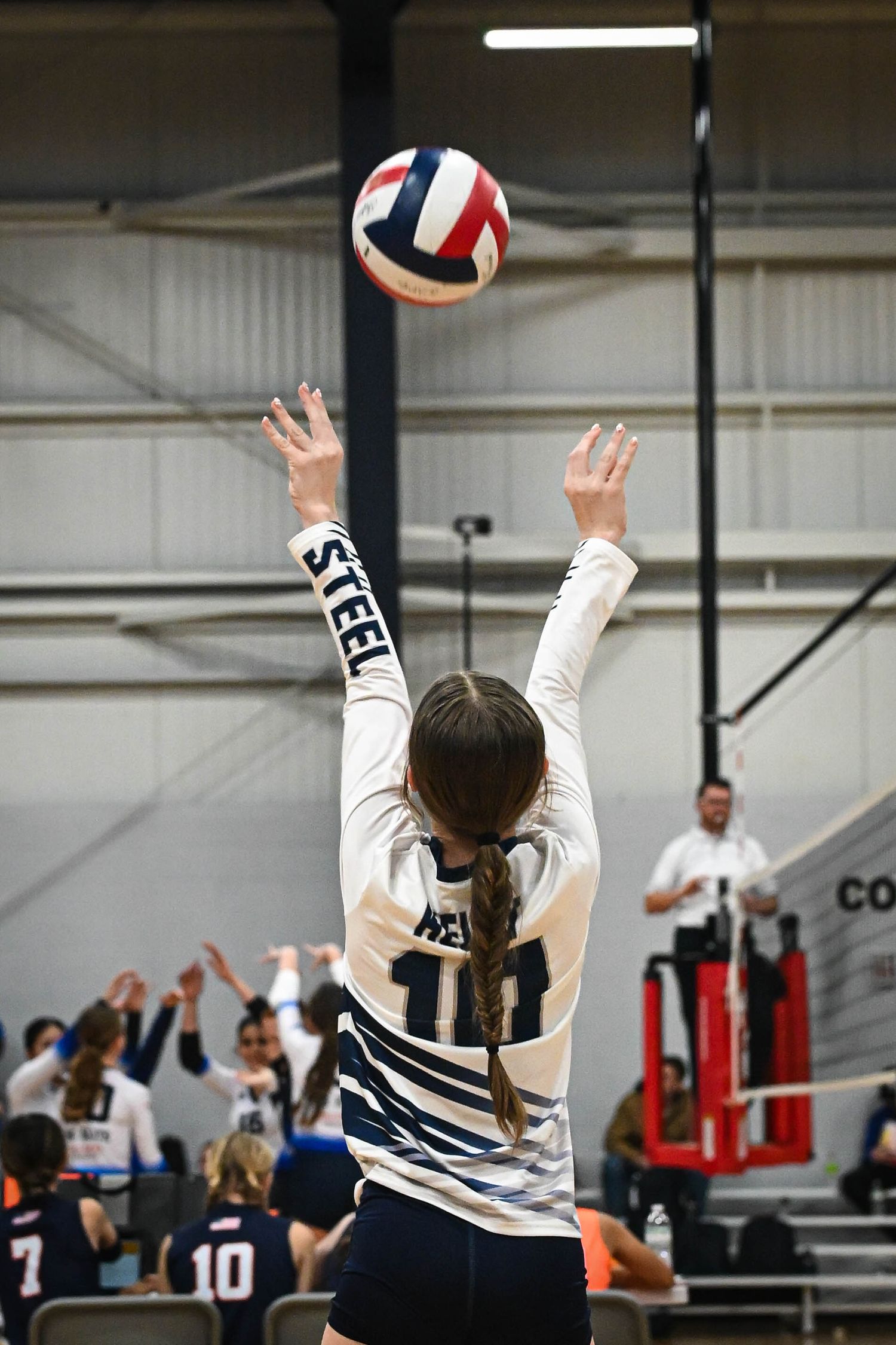 Volleyball player in white and navy uniform sets the ball during an indoor match against red and white team.