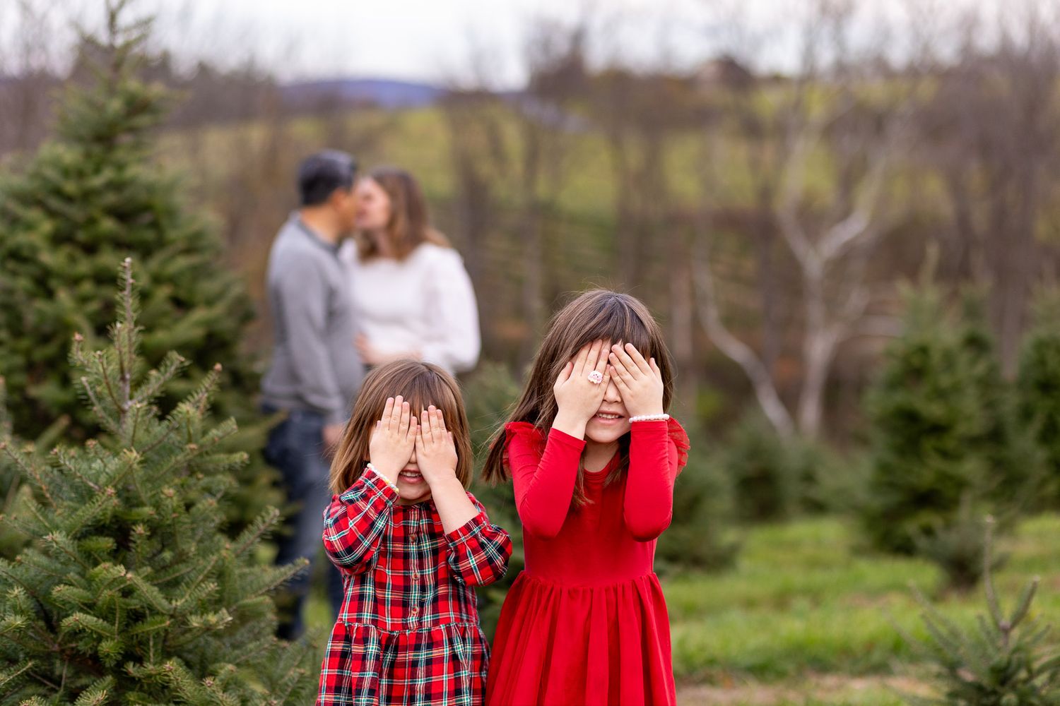 Two sisters in coordinating red and plaid outfits poses for outdoor winter photos among evergreen trees, their parents blurred behind.