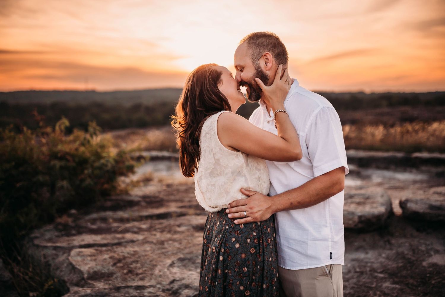 A couple shares a romantic moment together at sunset on rocky terrain while wearing casual attire.