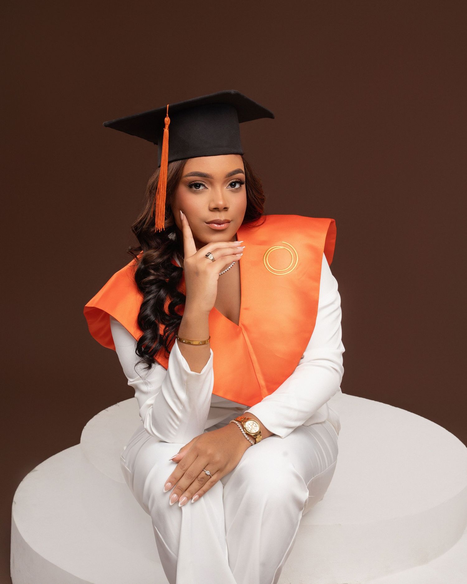 Graduate in black cap and gown poses with blue diploma holder in studio setting with brown backdrop.