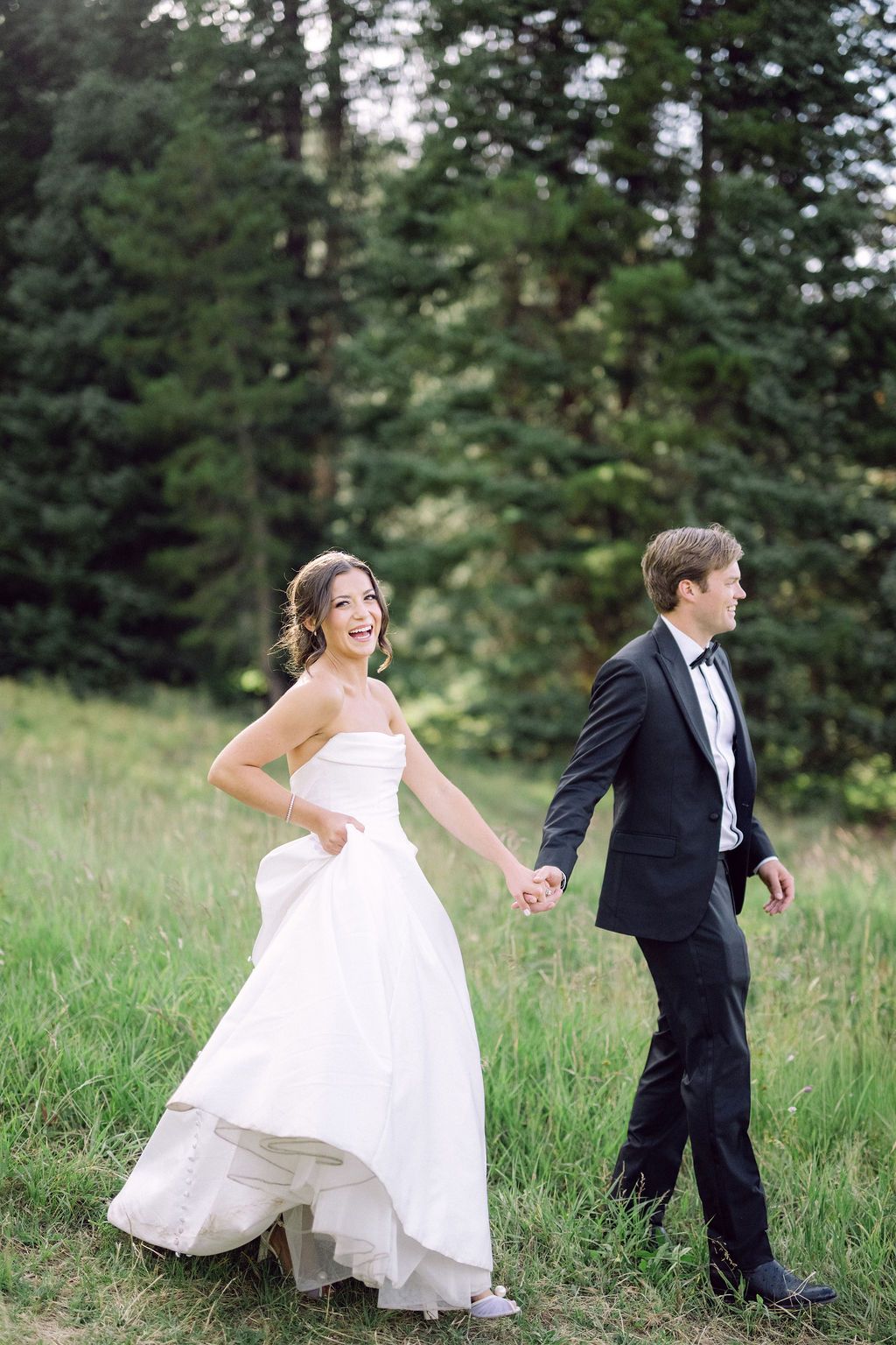 A bride in a flowing white gown and groom in black tuxedo pose playfully in a meadow with evergreen trees.