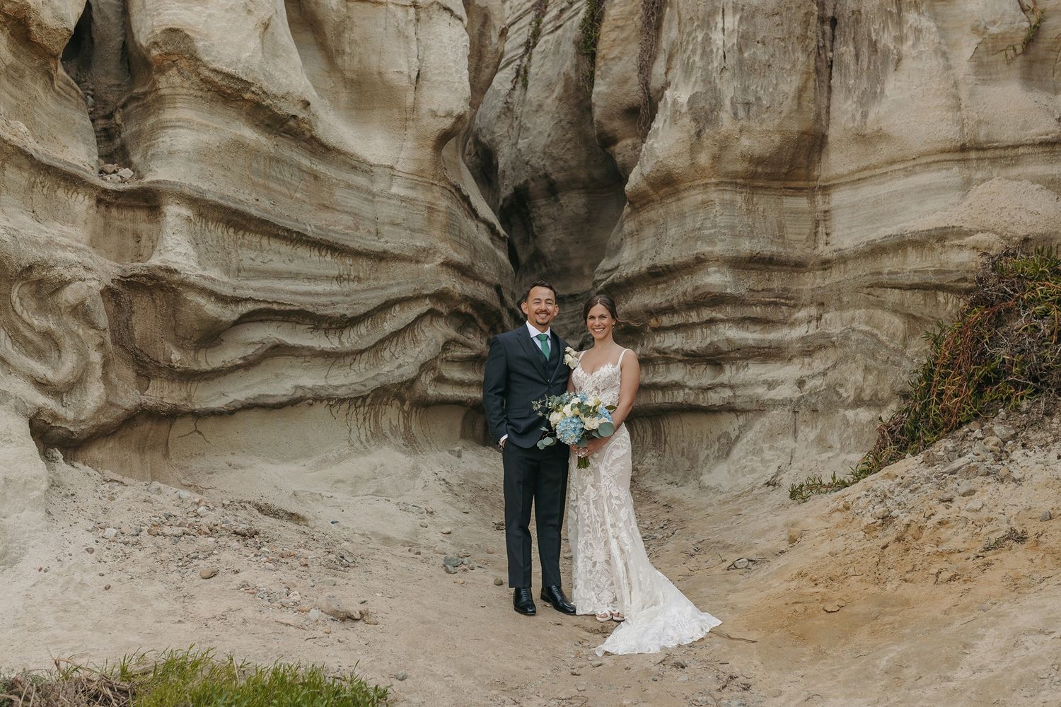 A couple in wedding attire stands in front of a rocky cliff formation with textured sandstone walls at Calafia Beach Park in San Clemente.