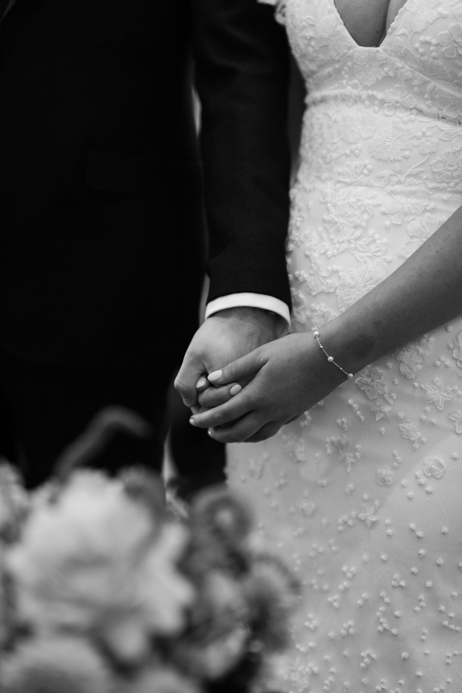 A close-up detail shot of hands holding each other against a white wedding dress with delicate beading and lace details.