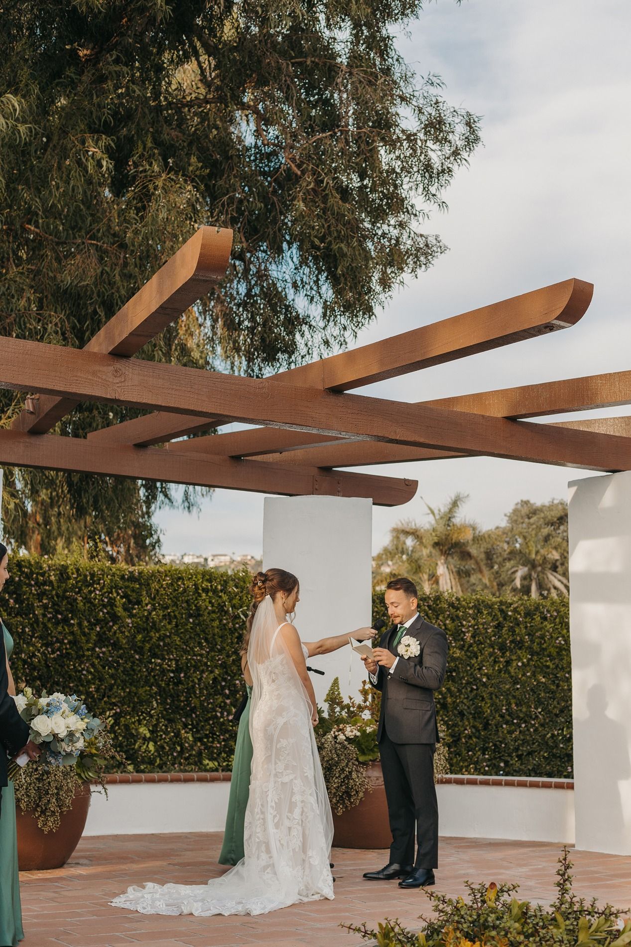 A wedding ceremony takes place under a wooden pergola with green hedges and trees in the background at San Clemente Shores.