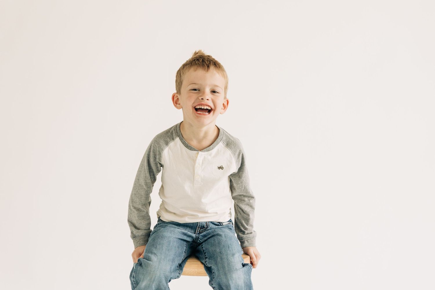 A child wearing a gray and white raglan shirt and jeans sits against a white background, smiling joyfully.