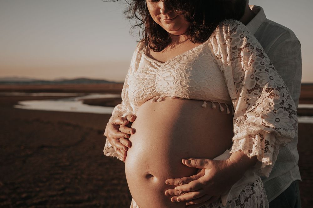 Intimate maternity photo at sunset showing pregnant belly in white lace dress with hands tenderly placed on bump.