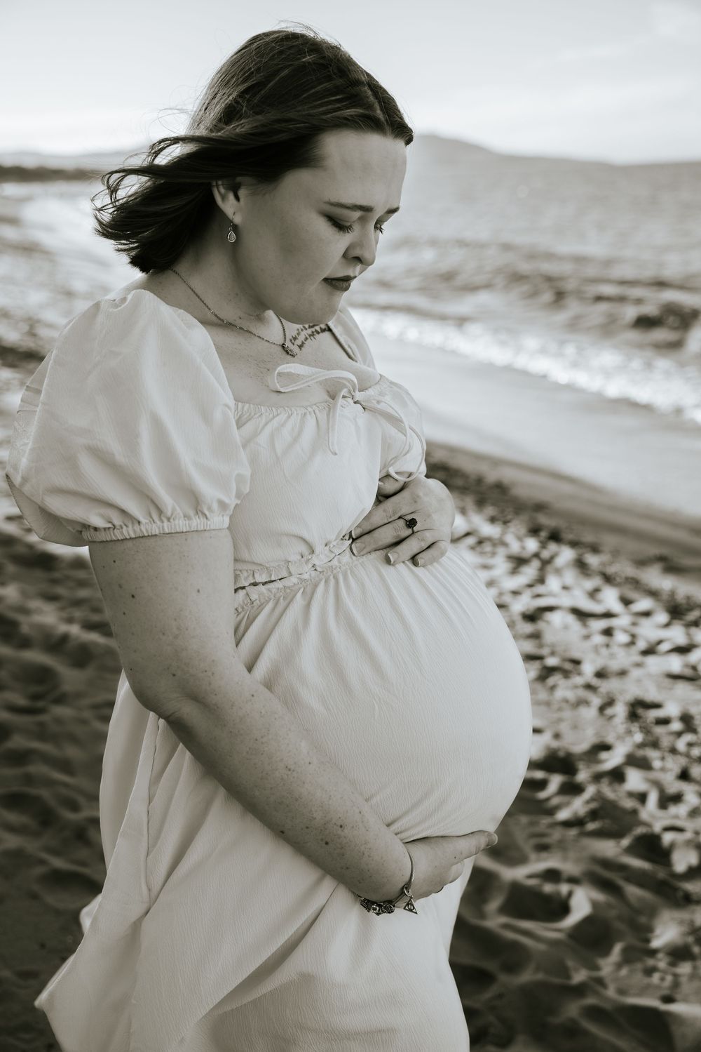 A pregnant woman in a white dress stands thoughtfully on a beach shore in black and white photography.
