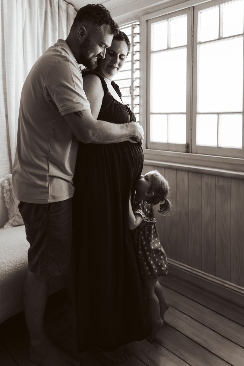Black and white maternity photo of couple embracing with toddler by window.