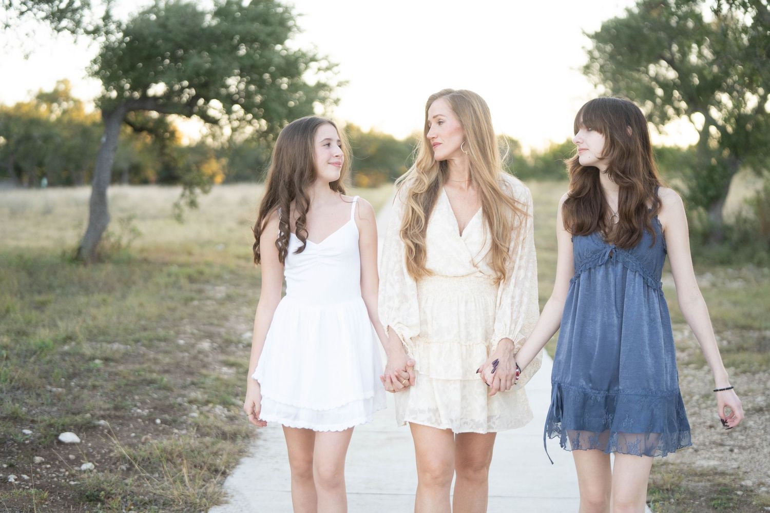 Three friends in white and blue summer dresses hold hands while walking and laughing together in a sunlit outdoor setting.