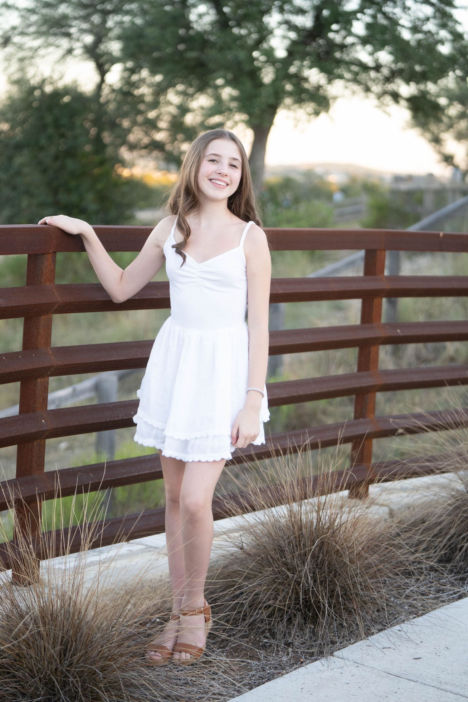A person in a white dress poses along a wooden railing with ornamental grasses at sunset.