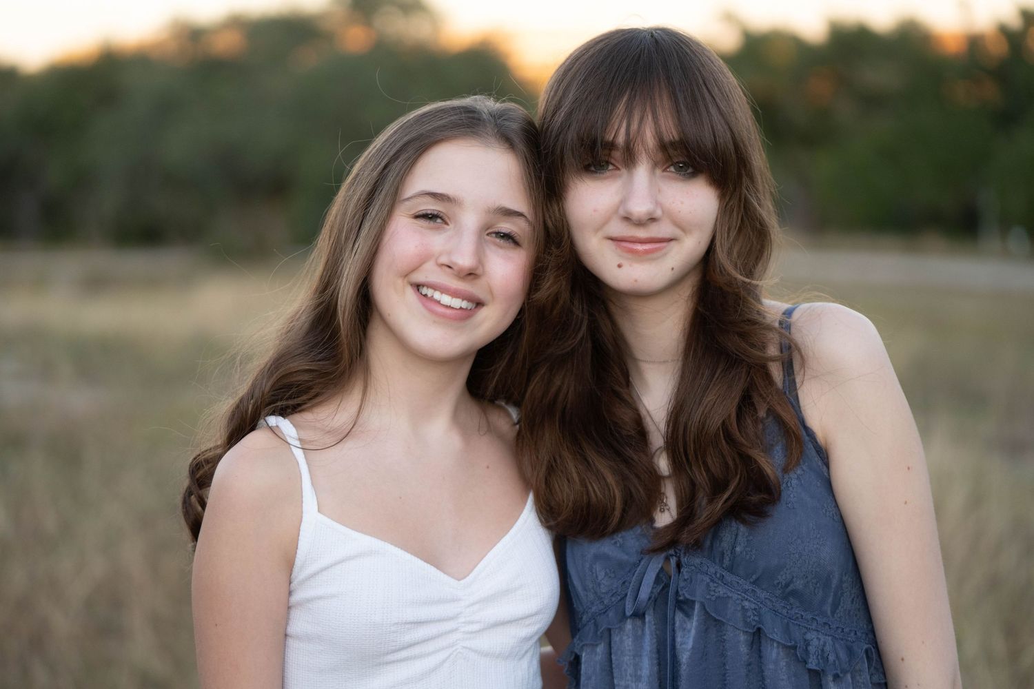 Two friends in casual summer outfits smile together in a grassy field at sunset.