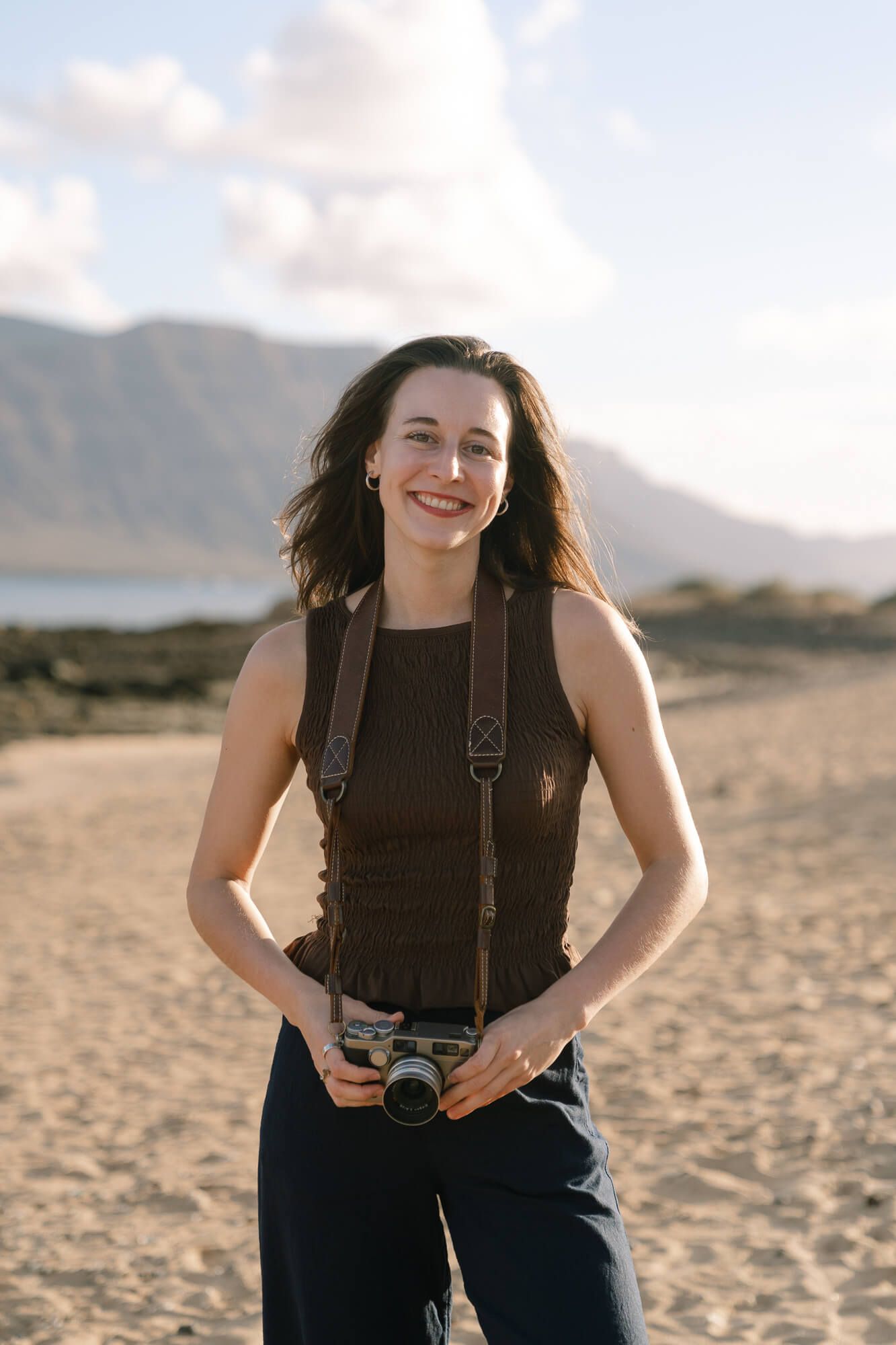 A photographer with a vintage film camera stands smiling on a sandy beach with mountains in the background.