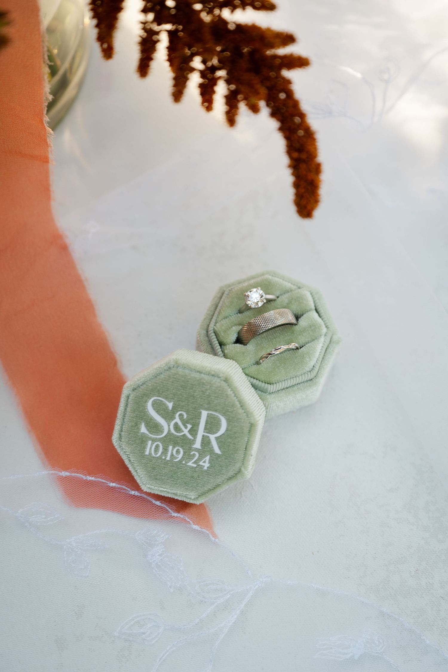 Sage green hexagonal ring boxes with initials displayed on white fabric under dried floral stems.
