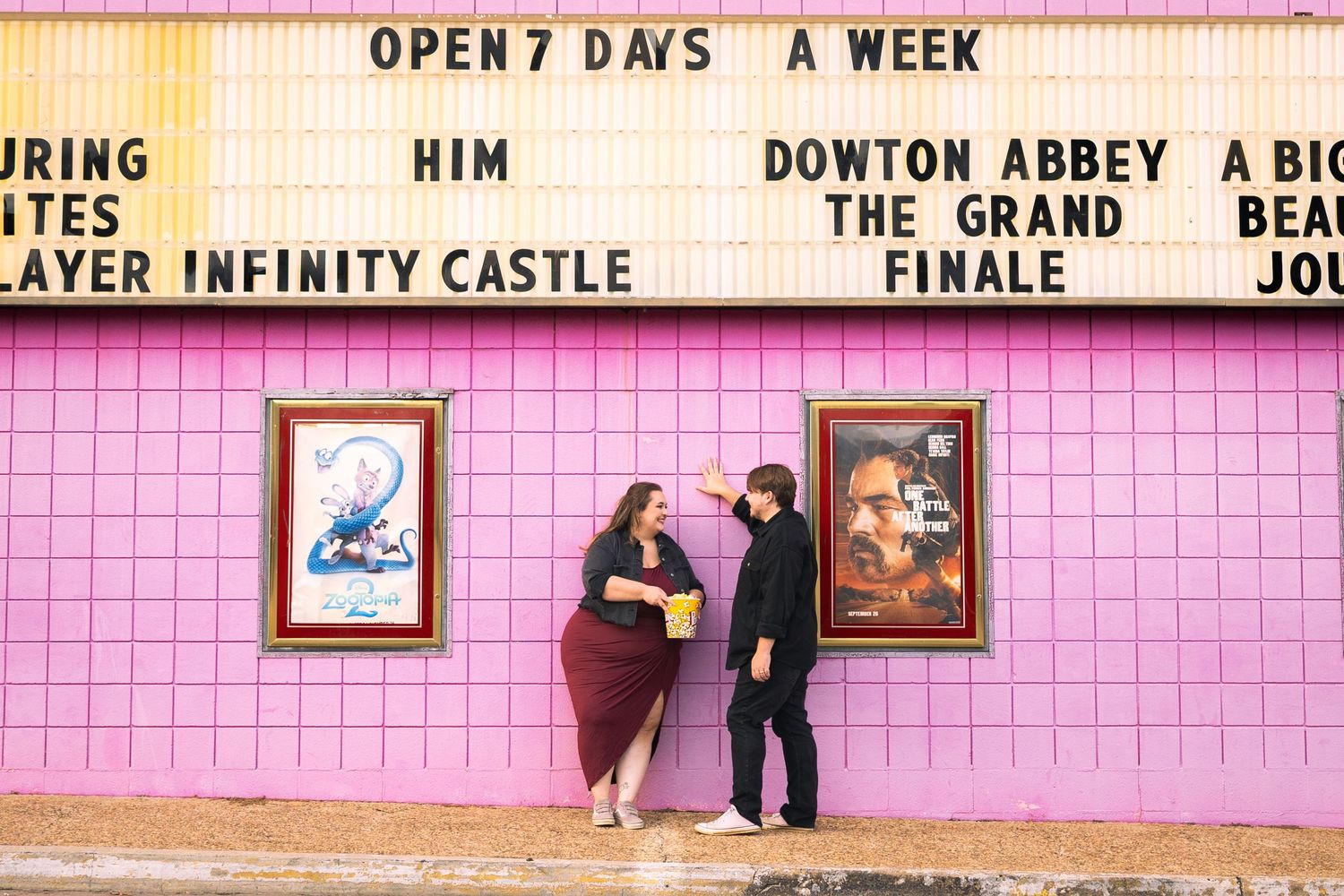 Two people stand in front of a pink wall with movie posters and showtimes at a theater entrance.
