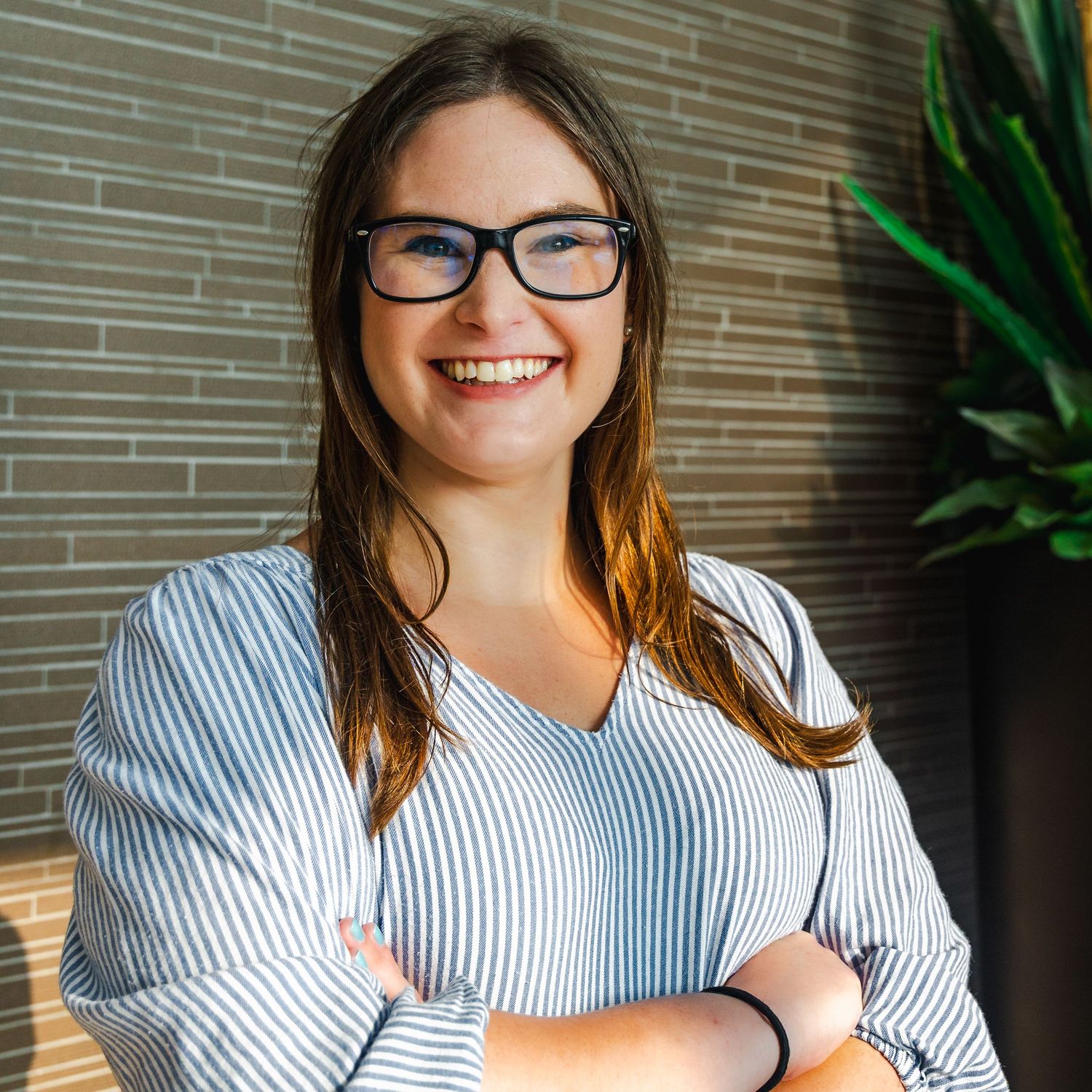 Professional portrait of someone in glasses and a striped top smiling warmly against a modern office wall with plants.
