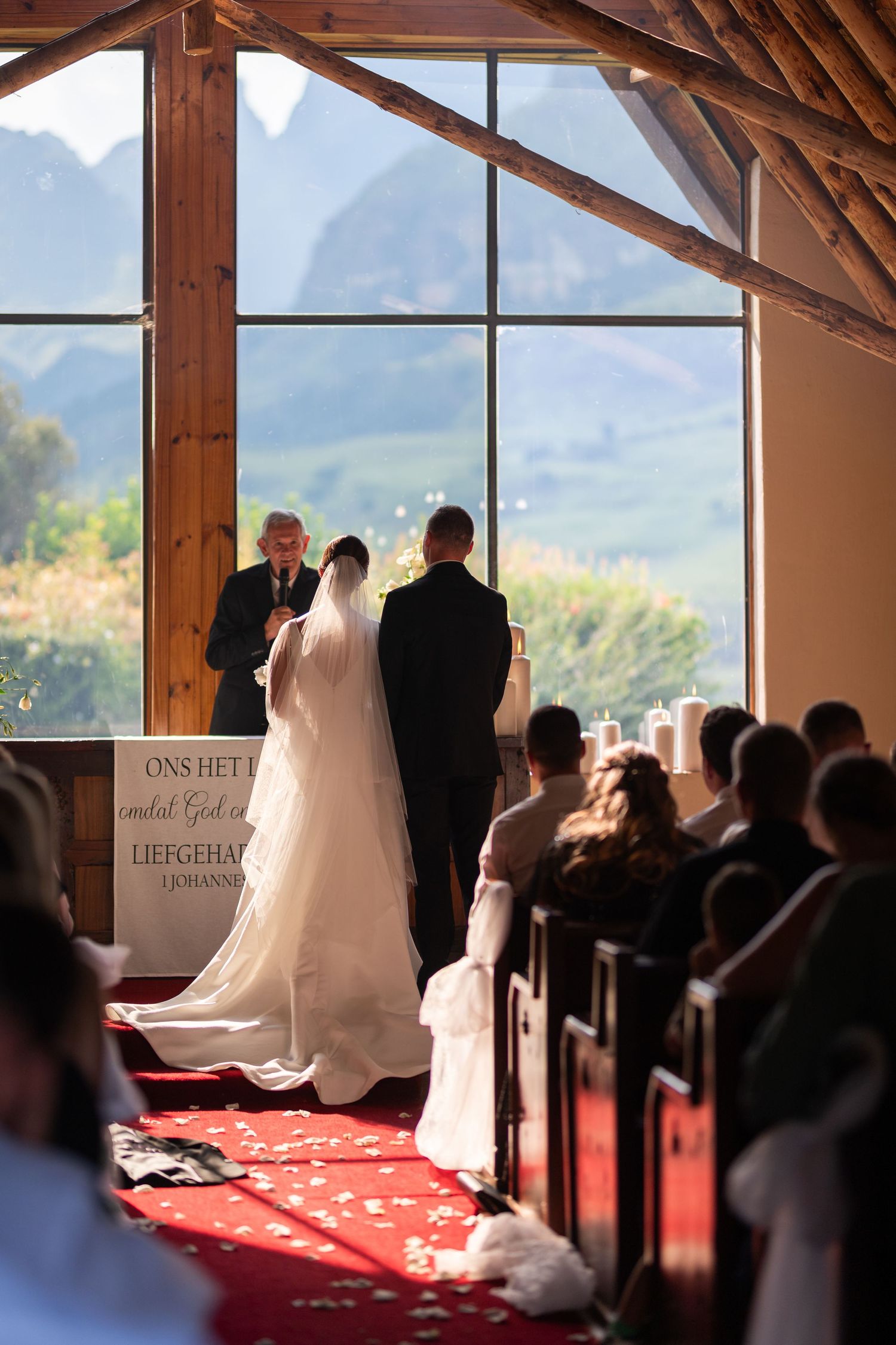 A wedding ceremony in a chapel with mountain views, bride and groom at the altar with guests seated in wooden pews.