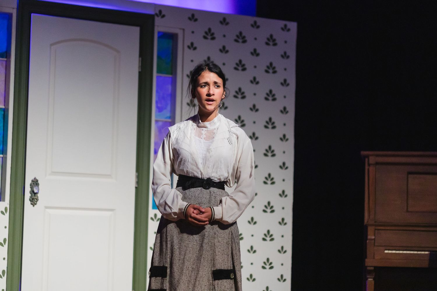 A person in a white shirt performs on stage with floral wallpaper and wooden furniture in the background.