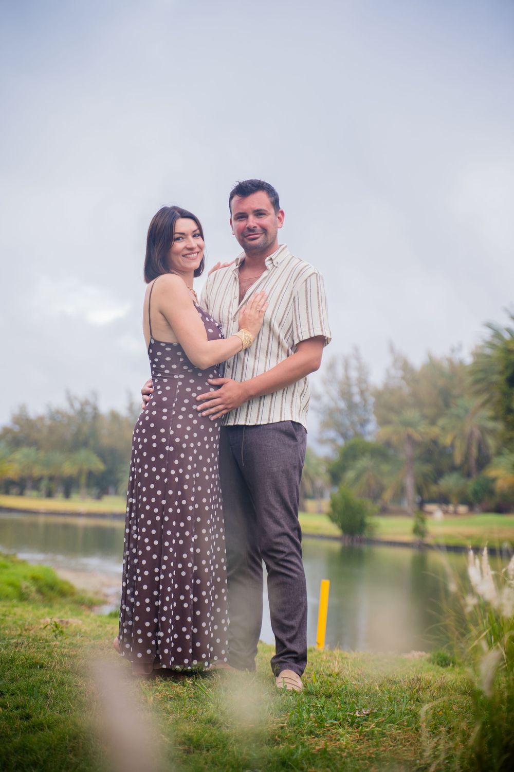 A romantic couple poses together by a serene lake surrounded by lush greenery.