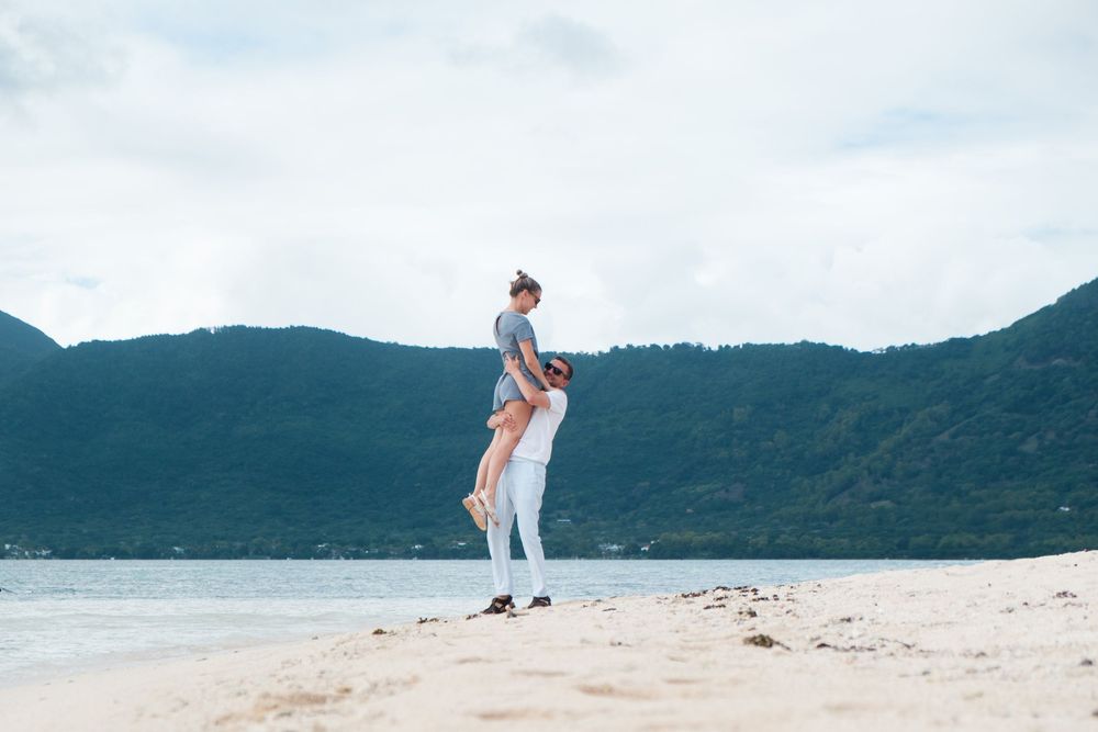 Couple walking hand in hand on a pristine white sand beach with mountains and ocean in the background.