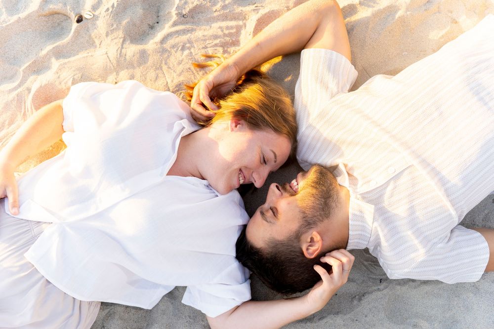 A couple lies together on white sand during a romantic beach photoshoot.