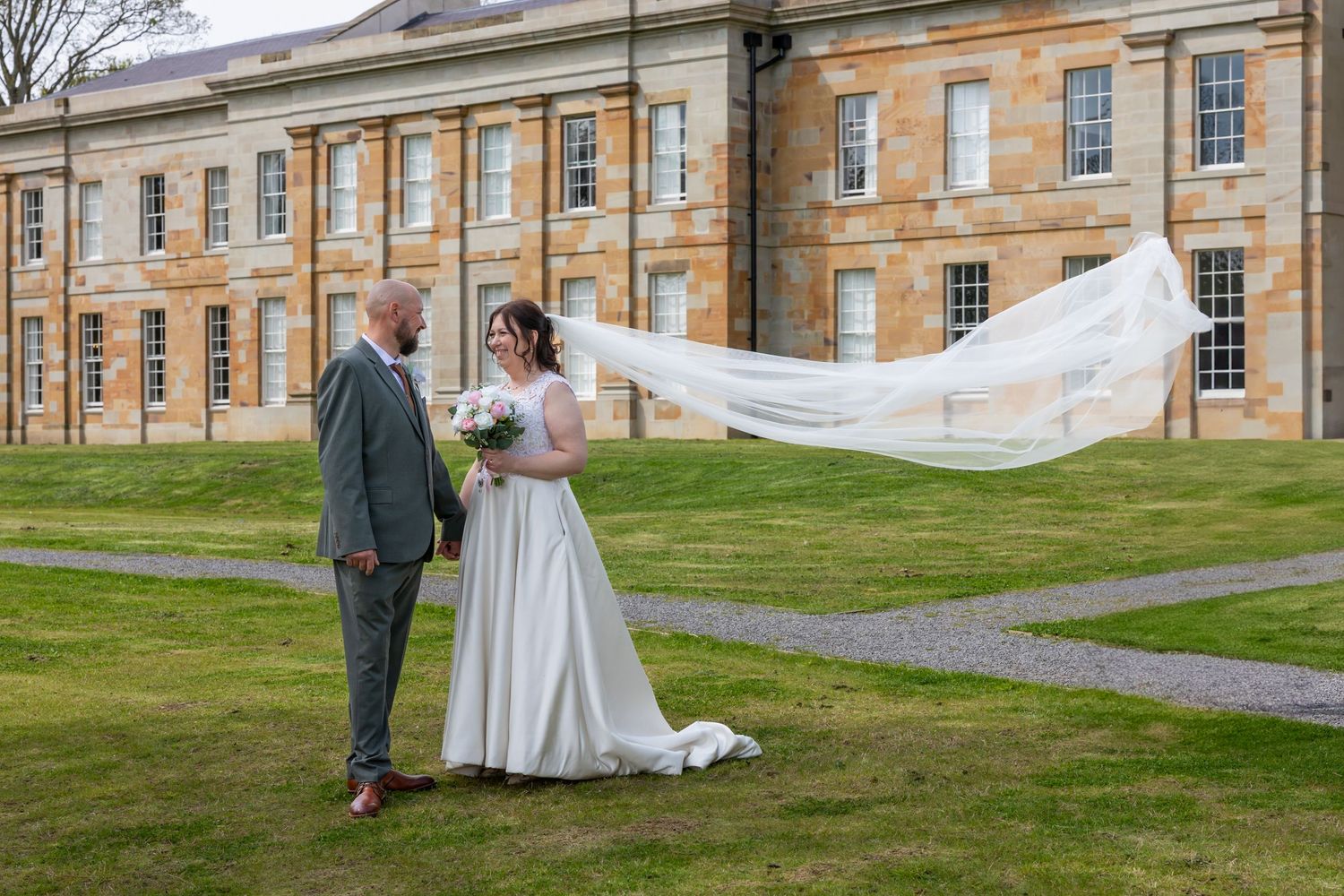 A bride and groom take wedding photos in front of a grand historic stone building on a grassy lawn. The Story Registry Office Durham