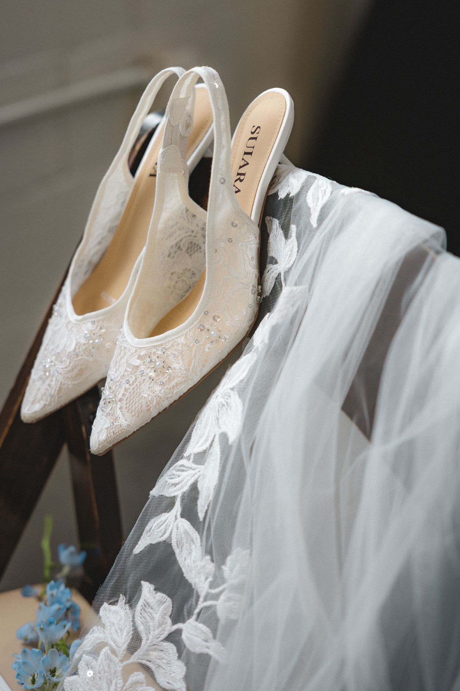 Elegant white lace bridal shoes displayed next to flowing tulle veil and delicate blue floral accents.