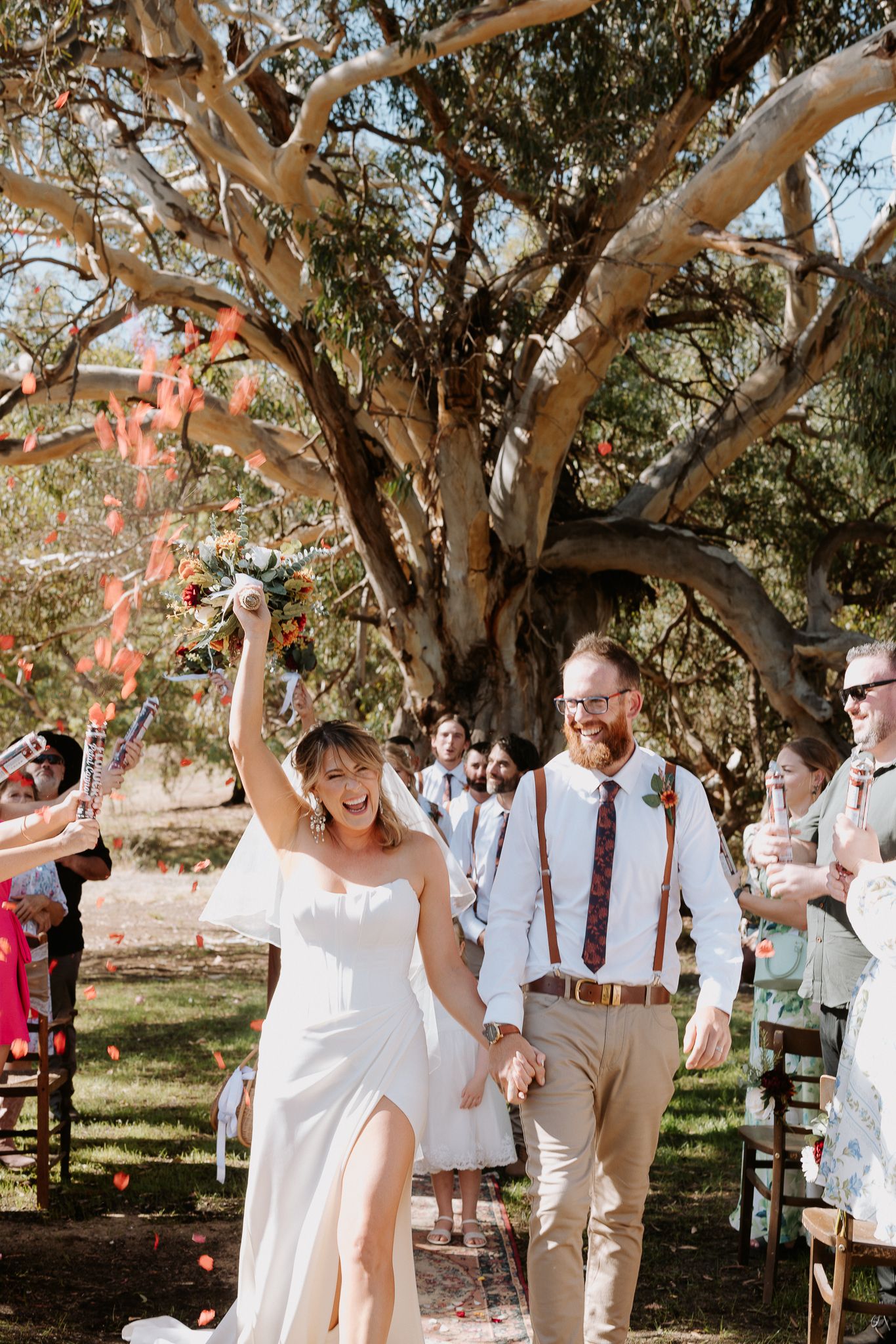 Outdoor wedding ceremony under large tree with guests seated on wooden chairs.