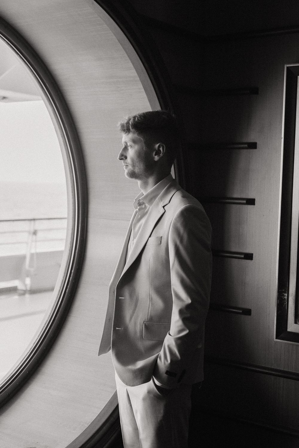 Elegant groom in a suit gazing through a circular porthole window in black and white.